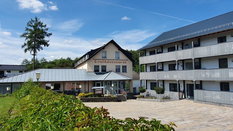 Ein modernes Gebäude mit dem Namen "EifelAdventures" in einer grünen Umgebung. Der Himmel ist klar und blau, was eine einladende Atmosphäre schafft.