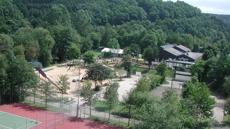 Freizeitpark mit Tennisplatz, Spielplatz und Restaurant in bewaldeter Umgebung. Im Hintergrund dichte Wälder, im Vordergrund Sport- und Spielanlagen.
