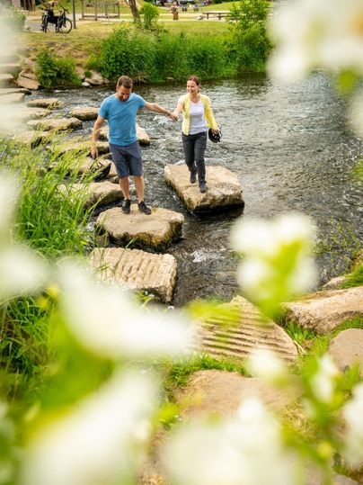 Ein Paar überquert auf Steinen einen Fluss im Kurpark Gerolstein. Im Vordergrund sind unscharfe Blumen, im Hintergrund grüne Wiesen und Bäume.