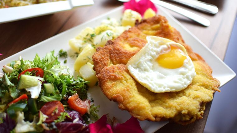 Ein Teller mit einem Schnitzel, darauf ein Spiegelei, daneben Kartoffeln und ein gemischter Salat. Im Hintergrund Besteck.