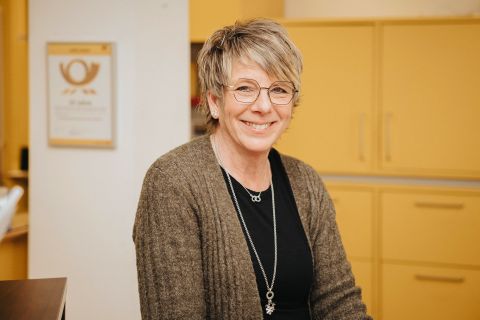 Eine Frau steht hinter einem Tresen eines Postschalters. Hinter ihr Schränke und das Posthorn-Logo.