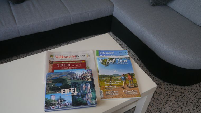 Eine helle Couch mit grauem Bezug steht neben einem weißen Tisch. Auf dem Tisch liegen mehrere Reiseführer über die Eifel und andere Reiseziele.