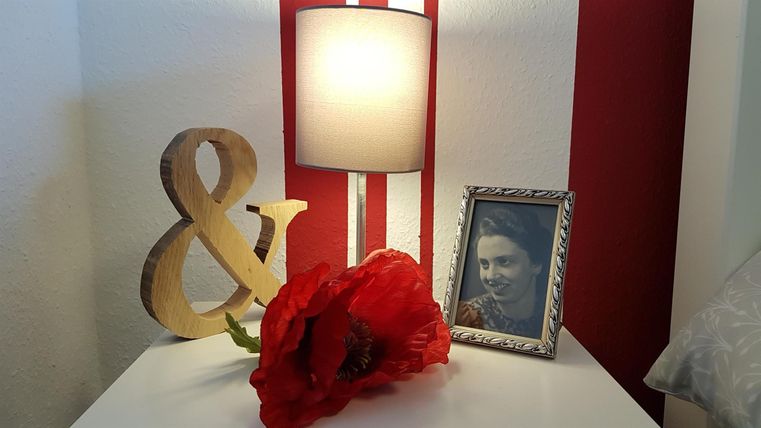 Ein stilvoller Tisch mit einer Lampe, einem Holzzeichen und einem roten Kunstblumenstrauß. Außerdem steht ein gerahmtes Bild auf dem Tisch.