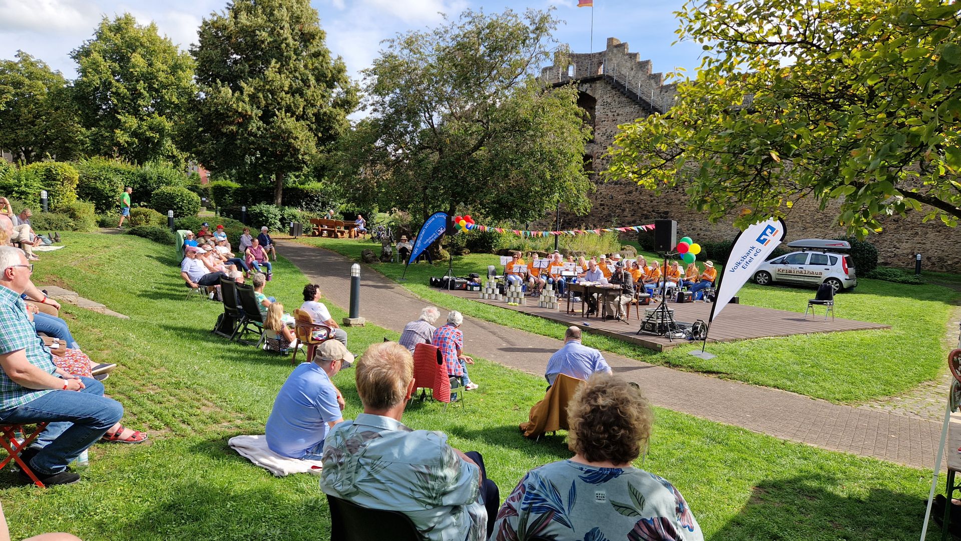 Zahlreiche Personen sitzen auf Stühlen und Decken auf einer Wiese im Park und schauen zur kleinen Bühne mit Musikverein und Lesern vor der Hillesheimer Stadtmauer