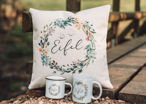 Handgemachtes Stoffkissen mit Blumemuster und Schriftzug "Eifel" in der Mitte auf einem Holzsteg. Davor zwei Tassen mit dem gleichen Design von Mitgebracht.