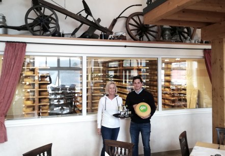 A man and a woman stand in front of a large glass pane, behind which numerous cheese wheels are maturing on shelves. The woman is carrying a tray with drinks and the man is carrying a cheese wheel.