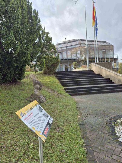 Ein modernes Gebäude mit Glasfassade und zwei Flaggen davor, umgeben von Bäumen und einem Schild im Vordergrund.