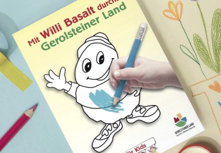 Voorpagina van het kleurboek “Met Willi Basalt door het Gerolsteiner Land” op een tafel vol kleurspullen en kleurrijke vellen papier.