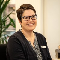 Eine Frau sitzt an einem Schreibtisch und lächelt in die Kamera. Hinter ihr ist eine große Palmenpflanze.