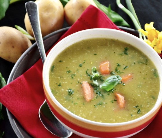 A bowl of potato soup on a saucer with a spoon, garnished with parsley, basil and carrot pieces. Next to it are potatoes and flowers.