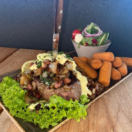 Ein Steak mit Zwiebeln und Sauce, garniert mit einem Messer, serviert mit Kroketten und einem frischen Salat auf einem Holztisch.