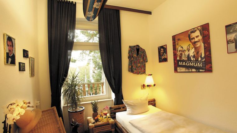 Une chambre confortable avec un lit simple et une décoration murale composée de photos et d'une planche de surf. Il y a une petite table avec une plante et des rideaux qui laissent passer beaucoup de lumière.