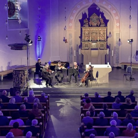 Fünf Musiker an Streichinstrumenten und Klavier musizieren vor dem Kirchenaltar. Davor die Sitzbänke voller Besucher.