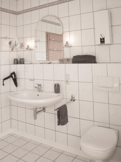 Ein modernes Badezimmer mit weißen Fliesen. Es enthält ein Waschbecken, eine Toilette und einen Spiegel.