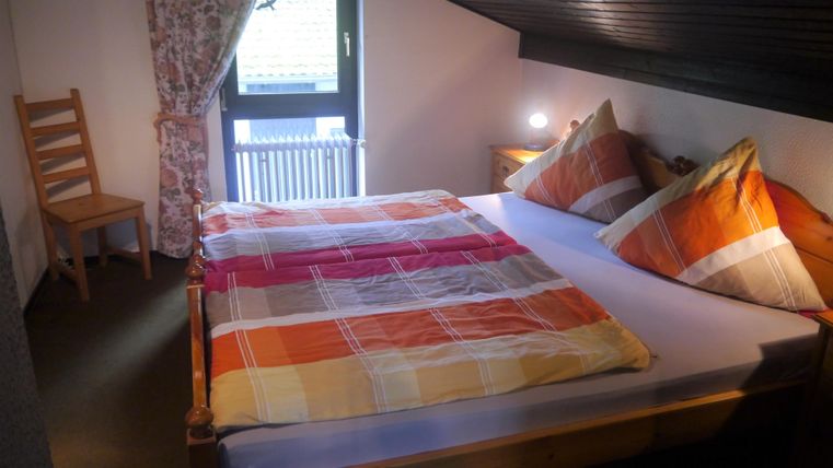 Ein gemütliches Schlafzimmer mit einem Doppelbett und bunten Kissen. Ein Stuhl und ein Fenster mit Vorhang ergänzen den Raum.