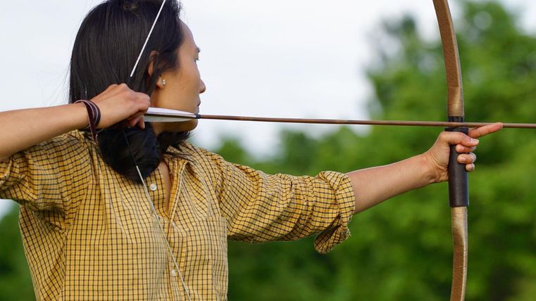 Eine Person in kariertem Hemd spannt einen Bogen im Freien. Der Fokus liegt auf der Haltung und dem Bogen.