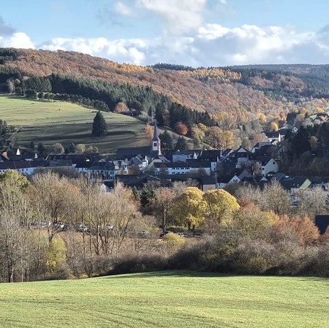 Panorama von Stadtkyll mit grünen Hügeln, herbstlichen Bäumen und einer Kirche im Vordergrund. Der Himmel ist blau mit einigen Wolken., © Touristik GmbH Gerolsteiner Land