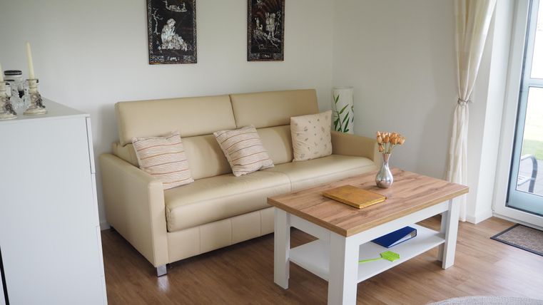 Ein helles Wohnzimmer mit einem beigen Sofa und einem Holzkaffee Tisch. An der Wand hängen zwei Bilder, und ein großer Fenster sorgt für viel natürliches Licht.