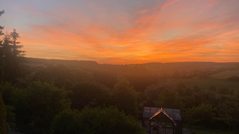 Ein atemberaubender Sonnenuntergang mit leuchtenden Farben über sanften Hügeln. Im Vordergrund steht ein kleiner Glashaus.