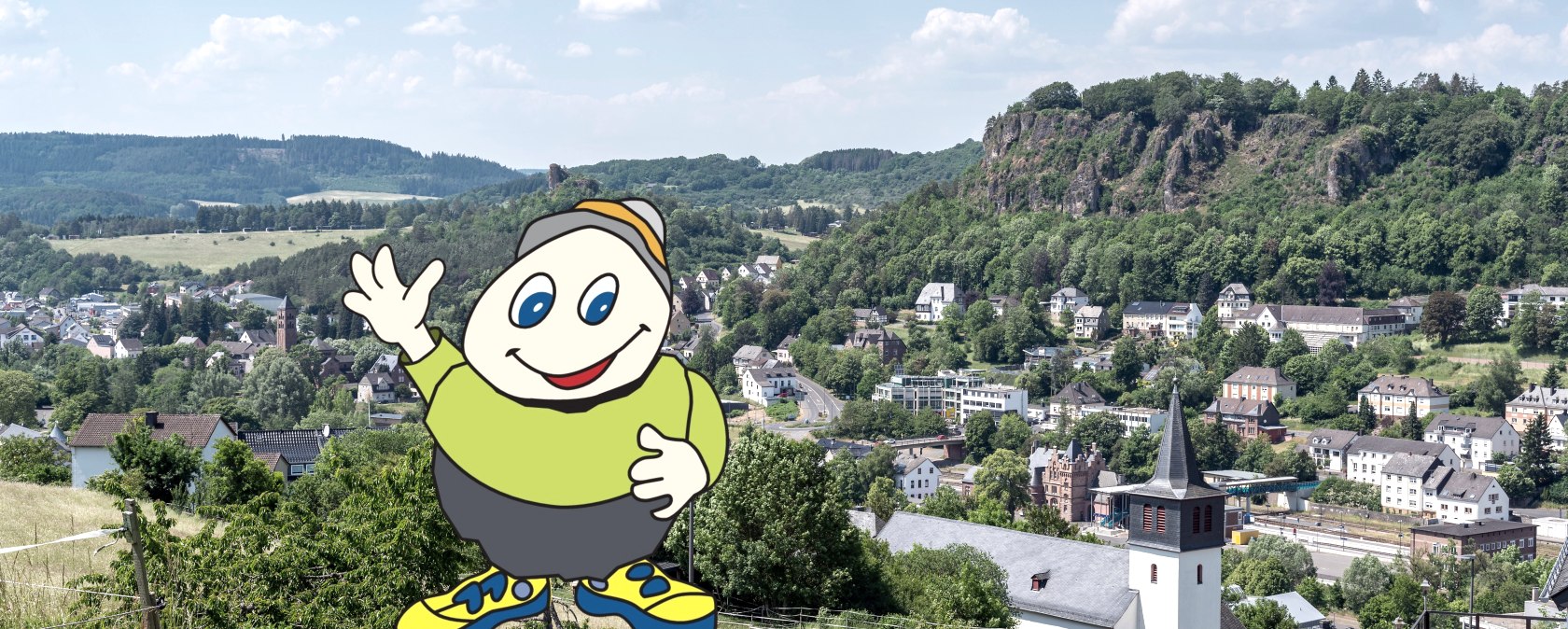 Cartoonfigur Willi Basalt mit grünem Shirt, grauer Hose und gelben Schuhen winkt. Hinter ihm Blick auf die Stadt Gerolstein mit Kirche, Gebäuden und Felsen.