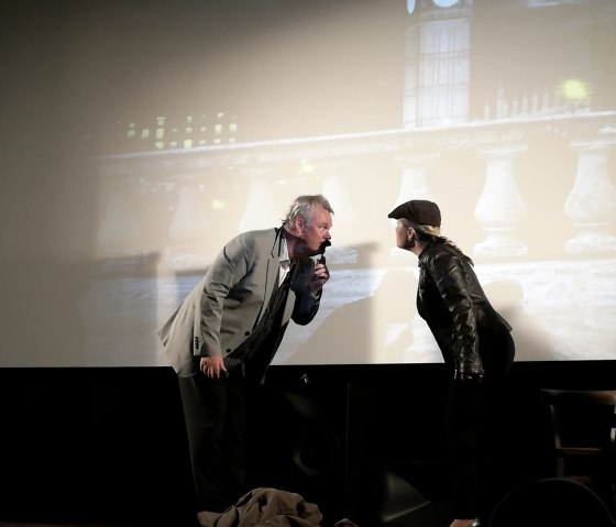 Zwei Schauspieler spielen eine Szene vor einer Kinoleinwand