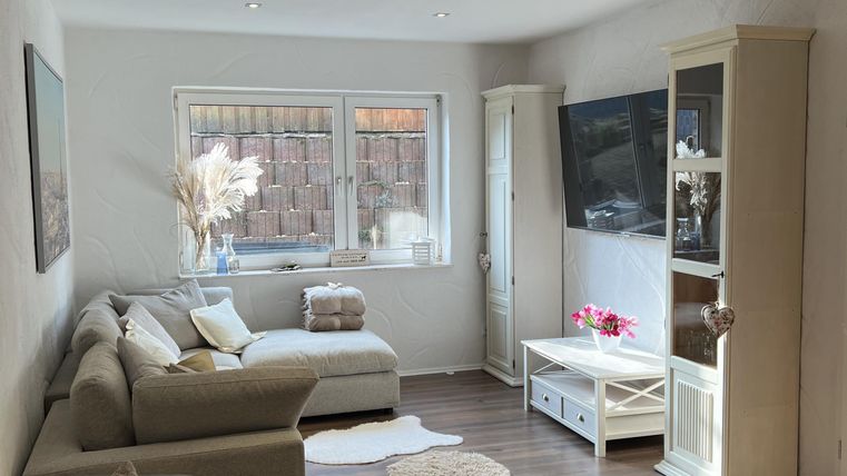 Een gezellige woonkamer met een lichte bank en een televisie aan de muur. Er is veel natuurlijk licht en een warme houten vloer.