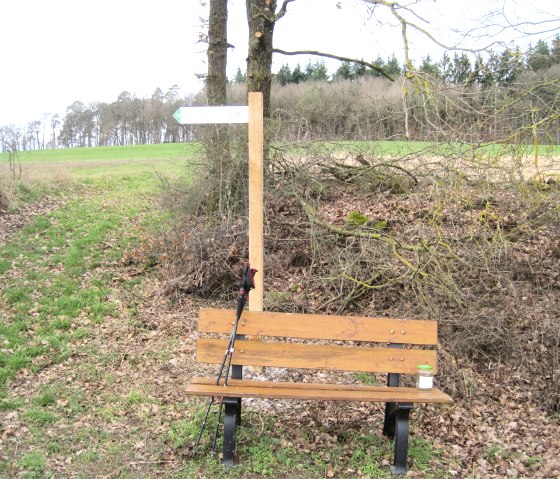 A wooden bench stands next to a signpost on a hiking trail. A walking stick leans against the bench, surrounded by trees and meadows., &copy; Touristik GmbH Gerolsteiner Land, Ute Klinkhammer
