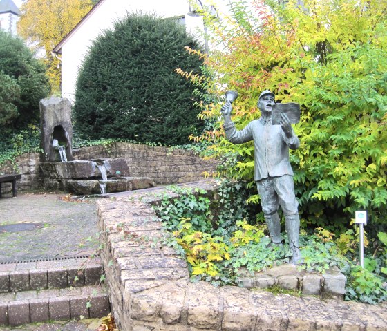 Eine Statue eines Mannes mit Glocke und Papier steht in einem Garten. Im Hintergrund sind ein Brunnen und dichte Büsche zu sehen., © Touristik GmbH Gerolsteiner Land, Ute Klinkhammer