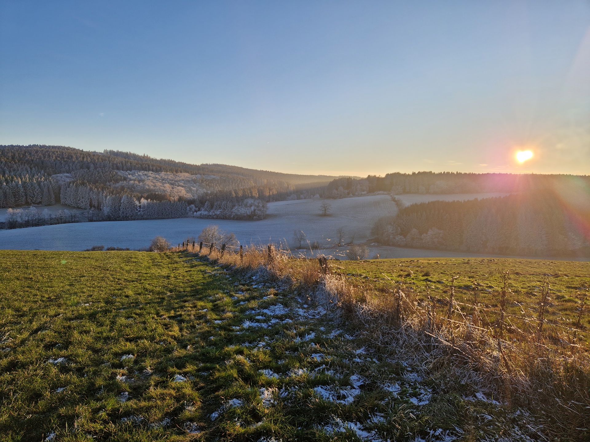 Hügelige Wiesenlandschaft leicht mit Schnee bedeckt und Sonnenuntergang im Hintergrund.