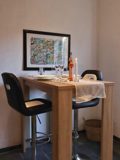 Een moderne eethoek met een hoge houten tafel en twee zwarte barkrukken. Op de tafel staan kaarsen, glazen en een fles wijn.