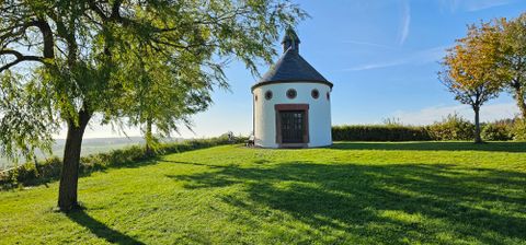 Eine kleine, runde Kapelle mit schwarzem Dach steht auf einer grünen Wiese, umgeben von Bäumen und blauem Himmel.