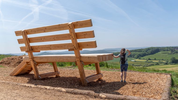 Eine riesige Holzbank steht in einer ländlichen Landschaft. Eine Person steht daneben und schaut in die Ferne.