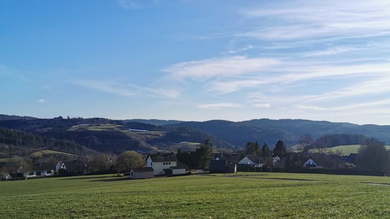 A green landscape with gentle hills and a clear blue sky. In the foreground, there are meadows and in the background, there are some houses.