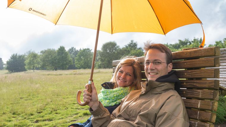 Un couple est assis sous un parapluie jaune sur un banc dans un paysage verdoyant. Ils sourient et profitent de l'atmosphère détendue.