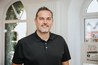 Ein Mann steht hinter einem Tresen und lächelt in die Kamera. Hinter ihr einige Fenster und Regale mit regionalen Produkten.