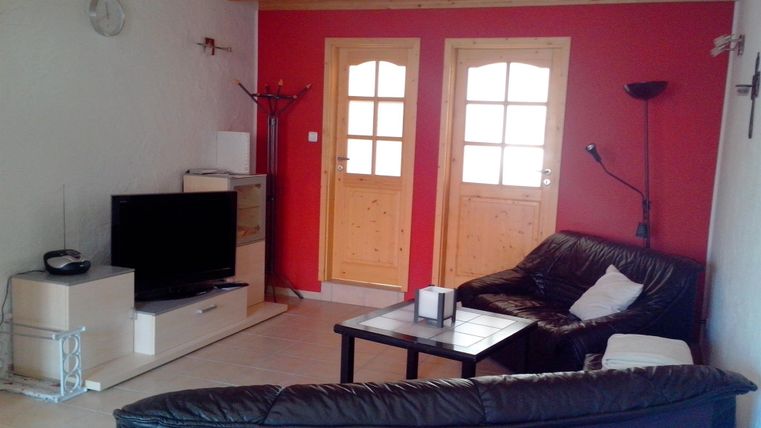 Ein gemütliches Wohnzimmer mit dunklem Sofa und einem Fernseher. Zwei Holztüren und eine rote Wand verleihen dem Raum eine warme Atmosphäre.