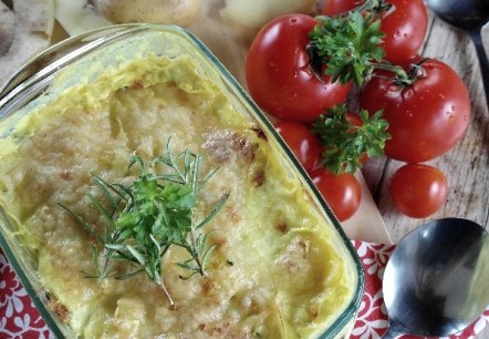 Eine Aufflaufform aus Glas mit einer mit Käse überbackenen Kartoffelzubereitung darin. Daneben Tomaten, Kartoffeln und Löffel.