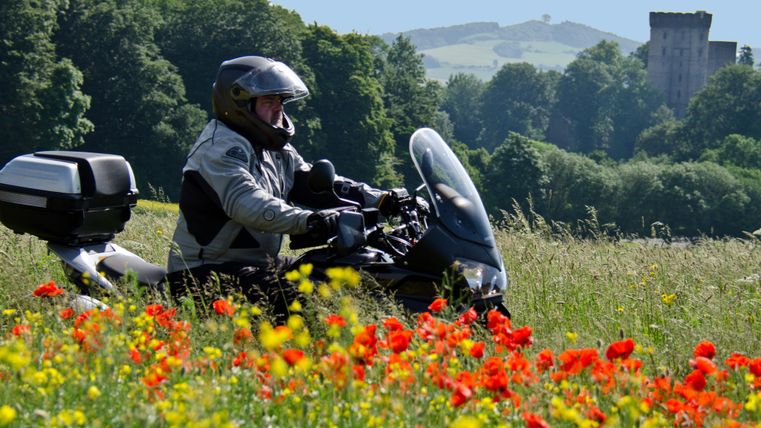 Un motocycliste se tient dans un champ de fleurs colorées. À l'arrière-plan, on peut voir des collines verdoyantes et une vieille ruine.