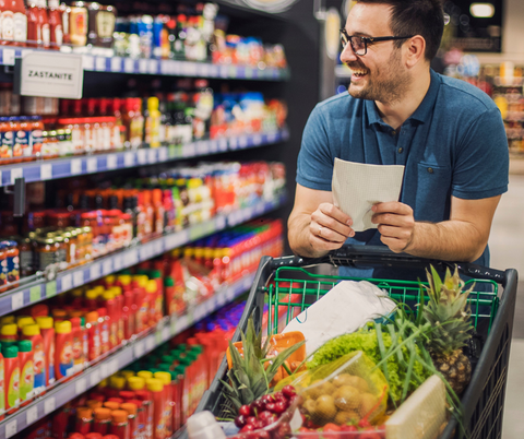 <p>Ein Mann mit einem gut gefüllten Einkaufswagen läuft durch einen Gang im Supermarkt. In den regalen sind Soßen und Ketchup zu sehen. In seiner Hand hält er eine Einkaufsliste.</p>