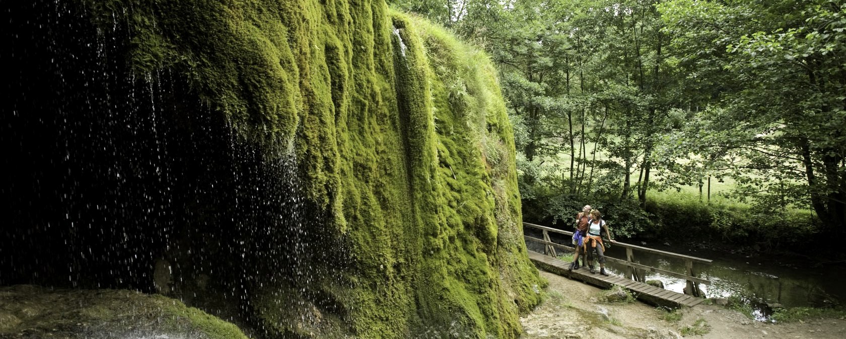 Der Nohner Wasserfall mit moosbewachsenen Felsen. Zwei Personen stehen auf einem Steg im Grünen. Ein idyllischer Zwischenstopp am Kalkeifel-Radweg., © Rheinland-Pfalz Tourismus GmbH/D. Ketz