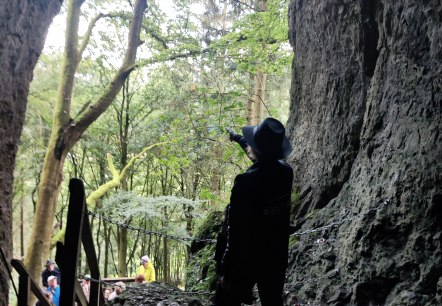 Steinernder Eingang in eine Höhle, in dem eine Person steht und eine Pistole auf den Wald vor der Höhle richtet. Ein Treppe führt hinauf zu Höhle und mehrere Personen stehen am Treppenanfang.