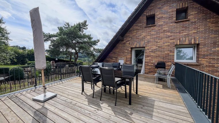 Eine sonnige Terrasse mit einem Esstisch und Stühlen. Im Hintergrund ist ein gepflegter Garten zu sehen.