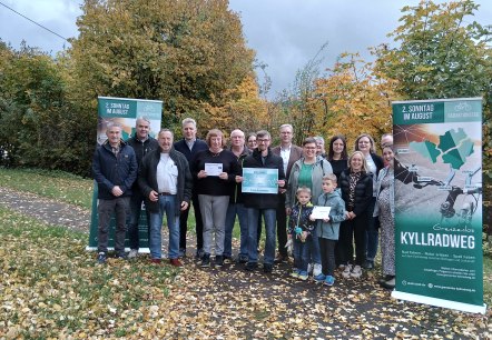 Eighteen people stand together in front of autumnal deciduous trees on a narrow asphalt road between two banners for the &lsquo;Grenzenlos Kyllradweg&rsquo; cycling event and smile for the camera. Three people proudly hold a prize in their hands.