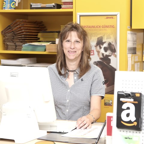 A woman stands at a post office counter and smiles at the camera. In front of her is a screen with a keyboard, and behind her are shelves full of envelopes, parcels and other postal items.
