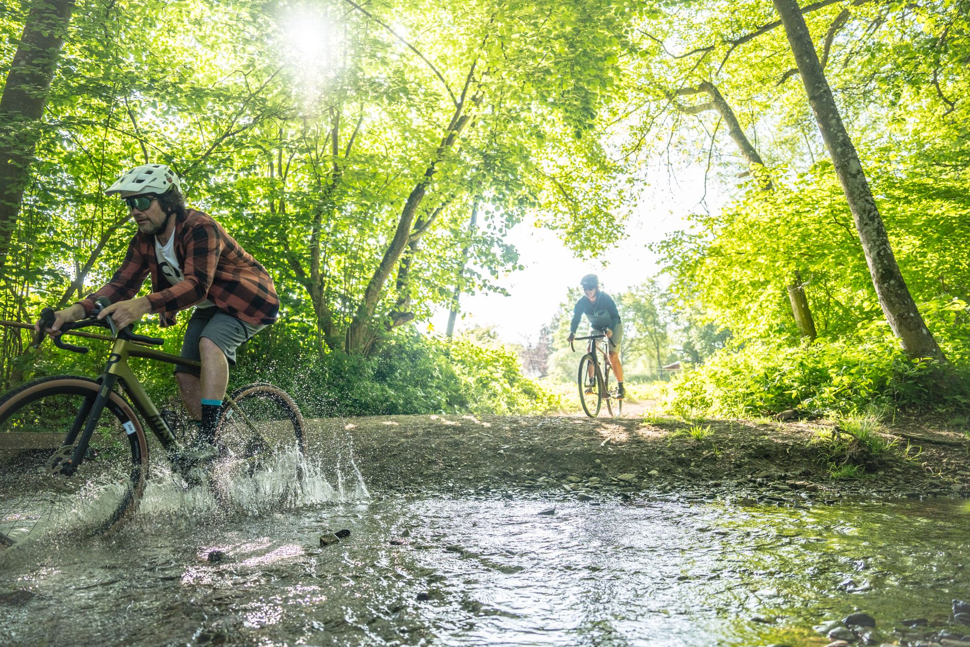 Ein Radfahrer auf einem Gravelbike fährt mitten durch eine große Fütze in einem Wald hindurch. Das Wasser spritzt zu allen Seiten. Hinter ihm fährt ein weiterer Radfahrer in den Wald hinein.