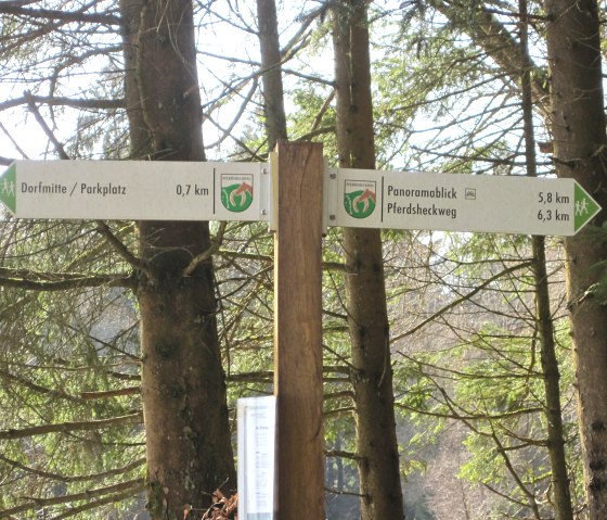 Signpost in the forest points towards the village center/parking lot (0.7 km) and Panoramablick Pferdsheckweg (5.8 km, 6.3 km). Surrounded by trees., &copy; Touristik GmbH Gerolsteiner Land, Ute Klinkhammer