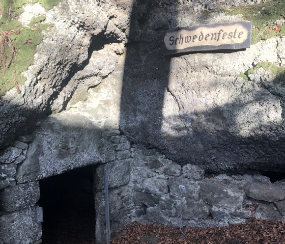 Ingang tot de Schwedenfeste in Hohenfels-Essingen, omgeven door rotsen en gebladerte. Een bord met de inscriptie 'Schwedenfeste' is zichtbaar., © Touristik GmbH Gerolsteiner Land