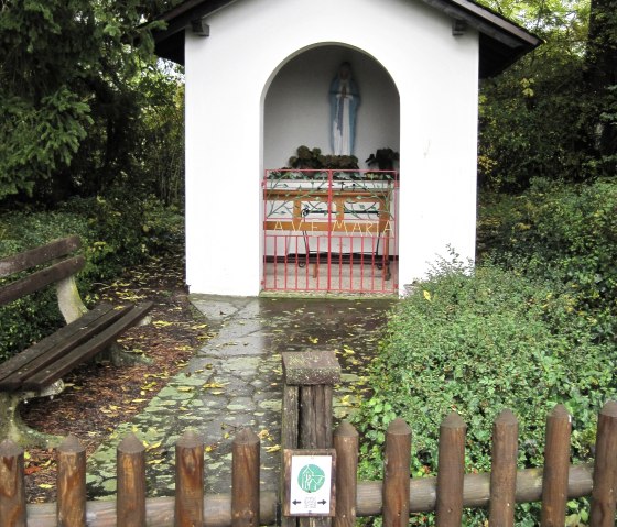 Kleine Kapelle mit Marienstatue, umgeben von Bäumen. Ein Holzzaun und eine Bank stehen davor. Herbstliche Blätter bedecken den Boden., © Touristik GmbH Gerolsteiner Land, Ute Klinkhammer