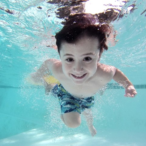 Ein kleiner Junge taucht unter Wasser in einem Schwimmbad und schaut lachend in die Kamera.