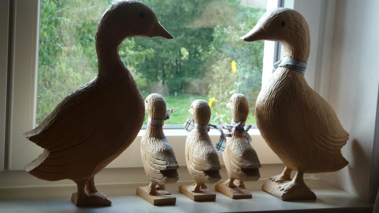 Wooden figures of ducks are standing on a windowsill. Two large ducks and two small chicks can be seen.